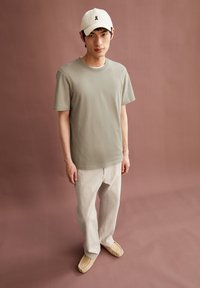 Junger Mann trägt eine beige Mütze, ein hellgraues T-Shirt, cremefarbene Hosen und beiges Slipper, während er vor einem gedämpften braunen Hintergrund steht.