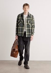 Chemise écossaise verte et noire avec deux poches poitrine, associée à un pantalon noir et un sac de voyage en cuir marron, portée avec des chaussures noires.
