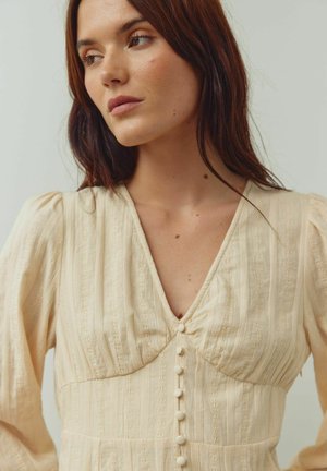 Krēmkrāsas kleita ar dziļu V-veida izgriezumu, teksturētu audumu, uzpūstām piedurknēm un dekoratīvām pogām priekšpusē. Smalkas vertikālas svītras.