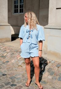 Femme aux longs cheveux blonds portant un haut bleu clair à manches courtes et un short assorti, debout sur un chemin pavé.