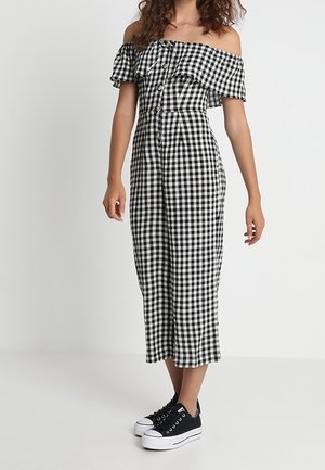 Kobieta ubrana w czarno-biały kombinezon w kratkę typu off-shoulder z guzikami z przodu i czarne sneakersy na platformie.