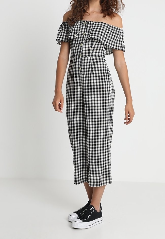 Femme portant une combinaison vichy noir et blanc épaules dénudées avec des boutons sur le devant et des baskets à plateforme noires.