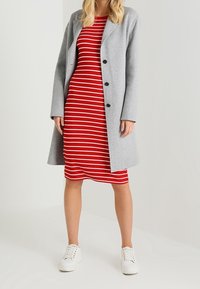 Harmaa pitkä takki mustilla napilla punavalkoisen raidallisen kapean mekon päällä, yhdistetty valkoisiin tennareihin. Sileä kangas; rento muotoilu.