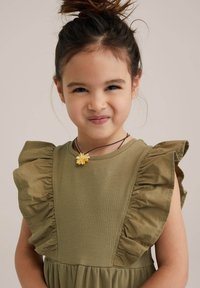 Olíva zöld ruha fodros ujjal, texturált anyagból, és redőzött szoknyával. Virág nyaklánccal díszítve, barna zsinóron.