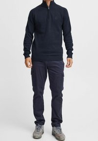 Pull à col zippé bleu marine en tricot côtelé, associé à un pantalon cargo foncé. Chaussures : baskets grises avec des accents rouges.