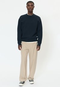 Felpa blu navy con girocollo, in misto cotone, vestibilità comoda. Pantaloni beige con finitura testurizzata, abbinati a scarpe da ginnastica nere.