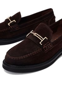 Mocasines de ante marrón oscuro con un diseño de costura elevada, que cuentan con un acento de hebilla metálica dorada en el empeine y una suela plana negra.