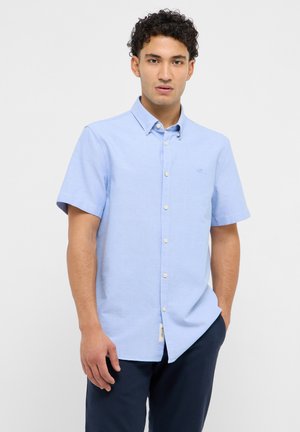 Mann trägt ein hellblaues Kurzarmhemd mit Knöpfen und dunkle Hosen, steht vor einem schlichten hellen Hintergrund mit einer Hand in der Hosentasche.