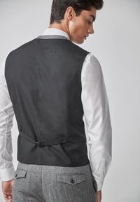 Schwarze Weste mit einem eleganten Design, kombiniert mit einem weißen Hemd. Auf der Rückseite befindet sich eine Schnalle und ein verstellbarer Riemen, dazu graue Hosen im Fischgrätmuster.