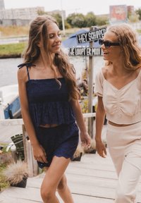 Twee personen lopen over een houten pad. De ene draagt een donkerblauwe, geknoopte crop top en shorts; de ander draagt een lichtbeige, geplooide crop top en broeken.