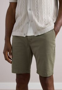 Lys beige strikjakke med korte ærmer og diamantmønster, lagdelt over en hvid ribbet skjorte, parret med olivengrønne shorts.