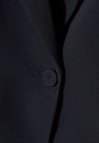 Gros plan sur l'avant d'un blazer noir montrant un bouton unique recouvert de tissu noir et un détail de revers.