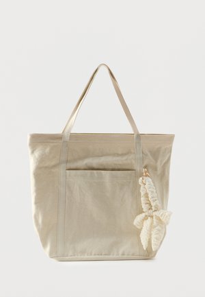 Sac fourre-tout en toile beige avec poche avant, longues poignées et un porte-clés scrunchie en tissu crème fixé sur le côté droit.
