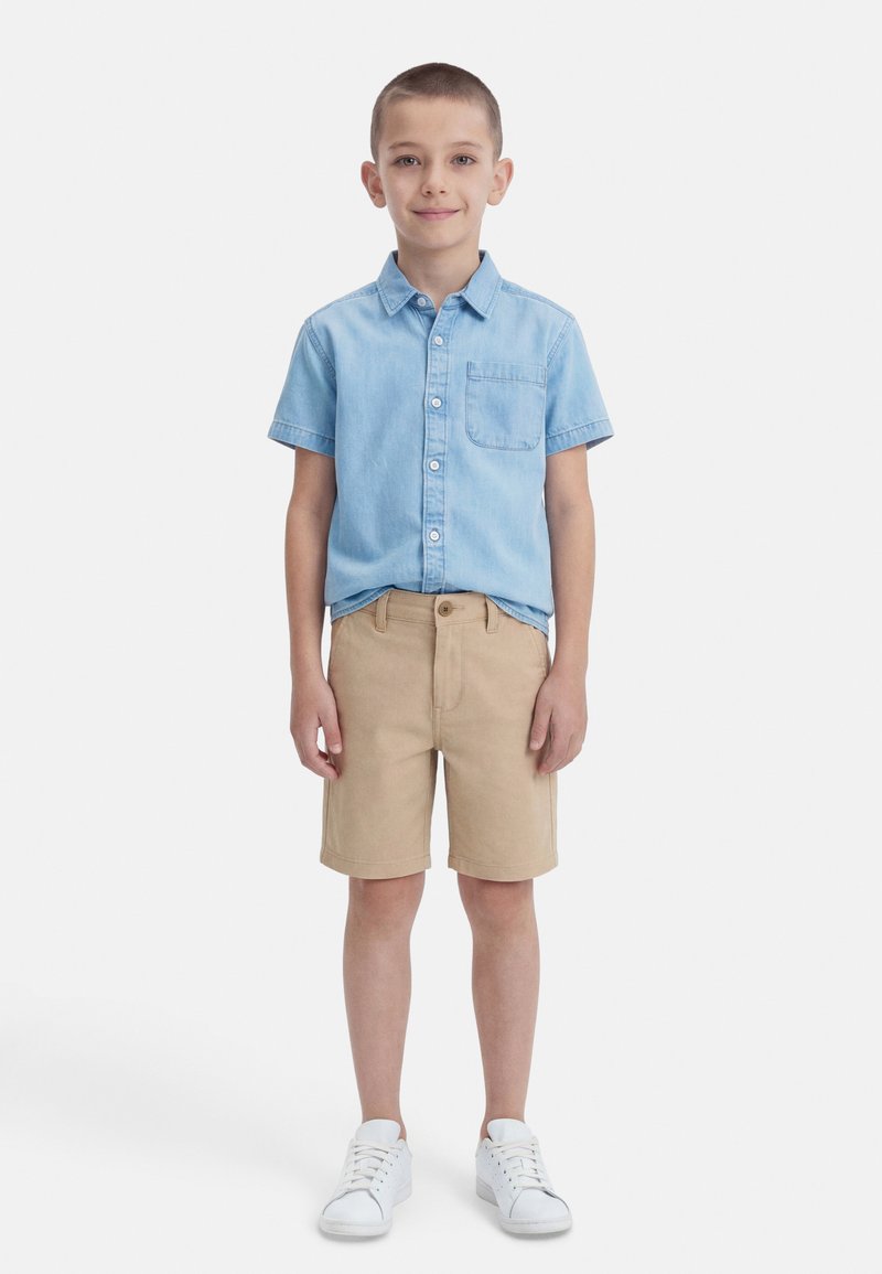 Αγόρι που στέκεται και χαμογελά, φορώντας ένα ανοιχτό μπλε πουκάμισο με κοντά μανίκια και κουμπιά, μπεζ σορτς μέχρι το γόνατο και λευκά αθλητικά παπούτσια.