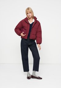 Chaqueta de plumas burdeos con un cuello alto, combinada con un suéter azul marino y jeans oscuros. El conjunto incluye zapatos planos granates brillantes y calcetines gris claro.