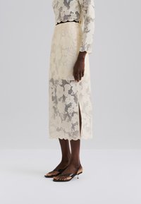 Spitzen-Midirock in hellem Gelb mit Blumenmustern, seitlichem Schlitz und gewelltem Saum. Kombiniert mit schwarzen minimalistischen Sandalen.