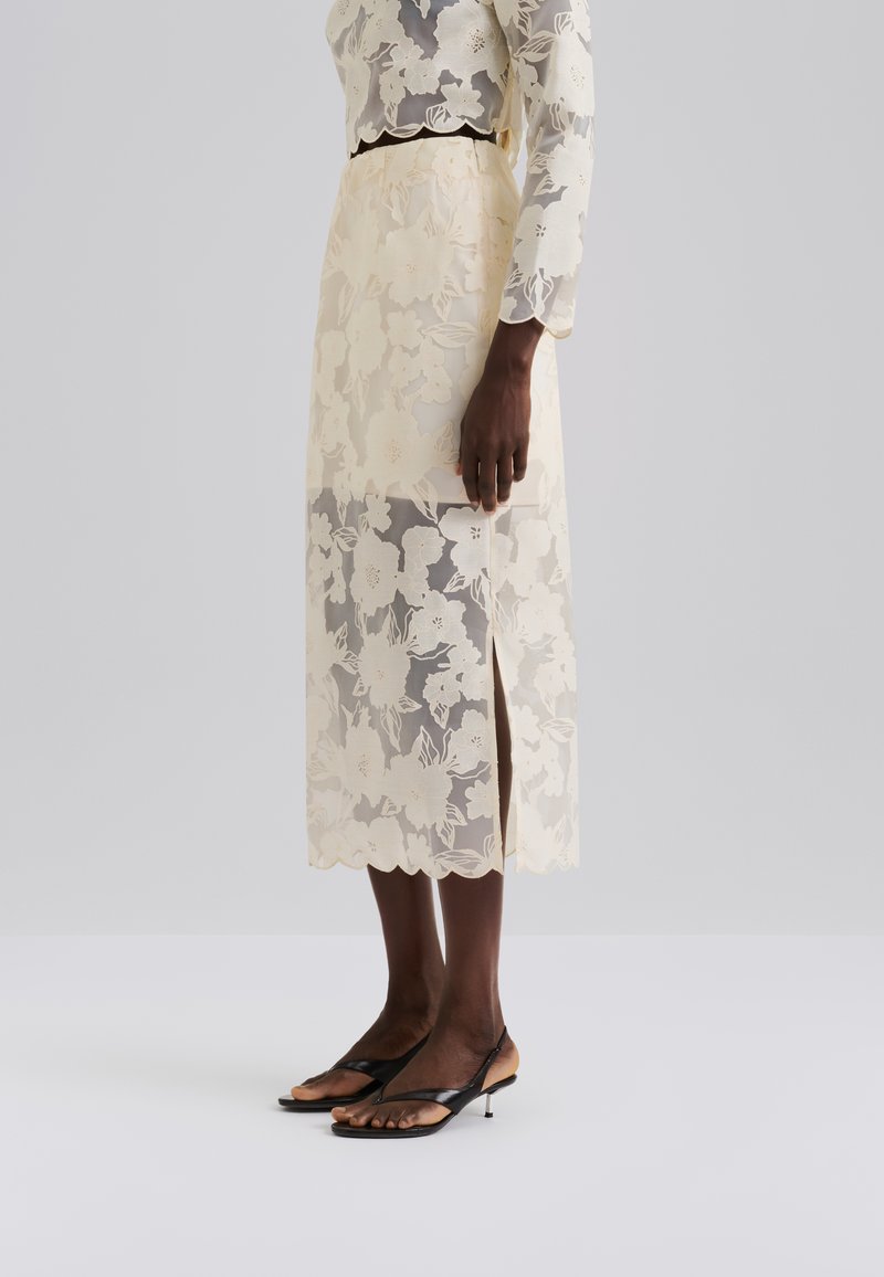 Spitzen-Midirock in hellem Gelb mit Blumenmustern, seitlichem Schlitz und gewelltem Saum. Kombiniert mit schwarzen minimalistischen Sandalen.