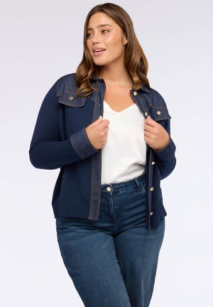 Eine Frau trägt eine marineblaue Strickjacke mit Denim-Details über einem weißen Oberteil und blauen Jeans. Sie hält die Strickjacke offen und schaut zur Seite.