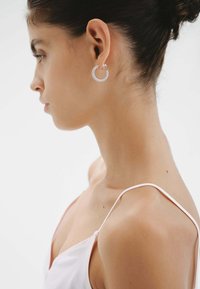 Pendientes de aro de plata con una superficie texturizada y delicados adornos, llevados en la oreja de una persona con un fondo neutro.
