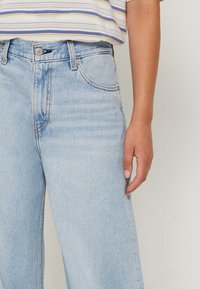 Svetle modré džíny s volným střihem, které mají pět kapes, zipový rozkrok a jemné vyblednutí na stehnech.