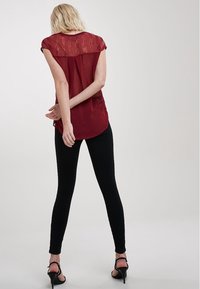Blusa marrón con espalda de encaje transparente, mangas cortas y dobladillo redondeado, combinada con leggings negros y sandalias de tacón alto negras.