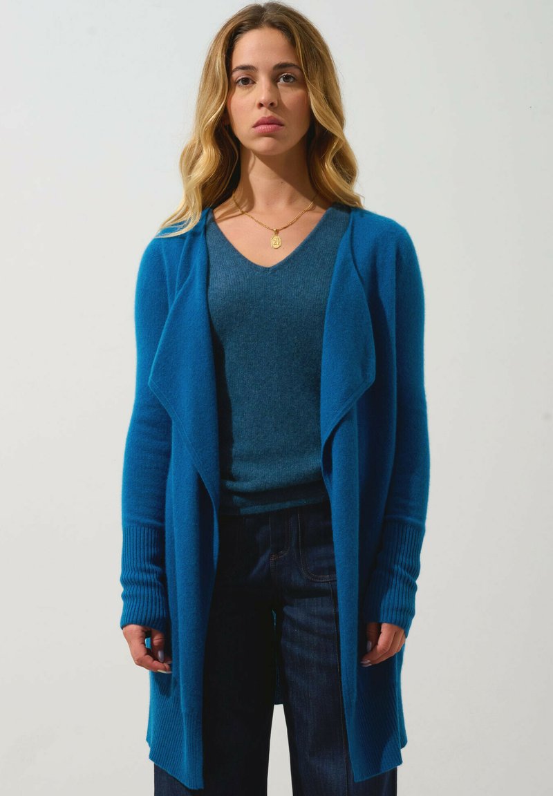 Cardigan lungo turchese con apertura frontale, polsini a coste e una tessitura morbida, indossato sopra una maglia blu a V coordinata e jeans scuri.