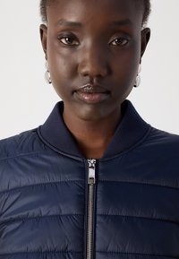 Laivastonsininen puuvillapufferitakki, jossa on vaakasuorat ompeleet, ribbireunus ja hopeaketju. Yhdistetty pyöreiden hopeakorvisten kanssa. Sileä, kiiltävä pinta.