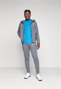 Grå Nike træningsdragt med hættetrøje og bukser, parret med en blå t-shirt. Hvide sneakers, glat stof og logo-detaljer synlige.