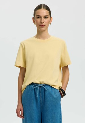 Vrouw met naar achteren gekamd haar die gouden creolen, een lichtgeel T-shirt, blauwe broek met trekkoord en armbanden om haar linkerpols draagt.