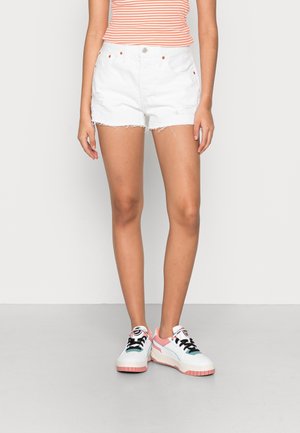 Femme portant un short en jean blanc effiloché, un haut rayé sans manches et des baskets blanches avec des détails roses et noirs, debout sur un sol gris.