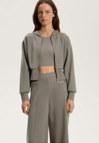 Giacca sportiva grigia con zip e cappuccio, design corto e polsini a costine, abbinata a pantaloni a vita alta coordinati e una maglietta aderente.
