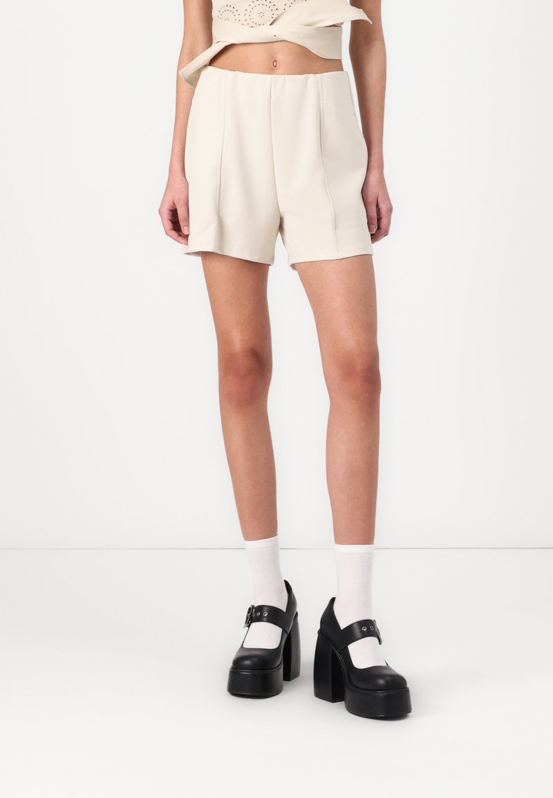 Shorts beige ajustés avec une taille élastique, ornés de deux plis à l'avant, associés à des chaussures à plateforme noires et des chaussettes blanches à la cheville.