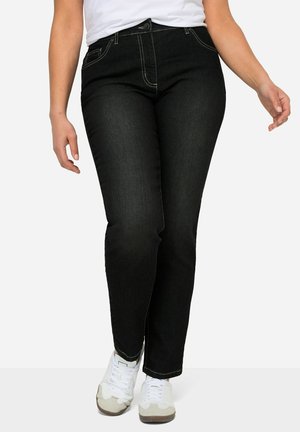 Personne portant un jean noir et des baskets blanches, marchant avec les mains détendues le long du corps, sur un fond blanc uni.