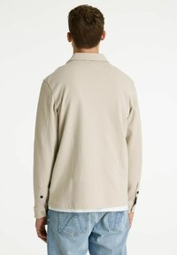 Camisa de manga larga de color beige claro con textura, con cuello, puños con botones y un ajuste relajado, llevada sobre pantalones vaqueros de mezclilla azul claro.