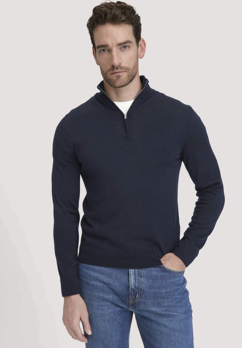 Marineblauer Reißverschluss-Pullover mit langen Ärmeln, gerippten Bündchen und Saum. Über einem weißen T-Shirt getragen, kombiniert mit blauen Jeans. Glatte Textur.