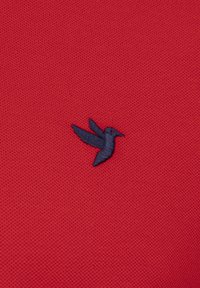 Tejido texturizado rojo con un pequeño logo de colibrí bordado en azul marino situado en el lado inferior.