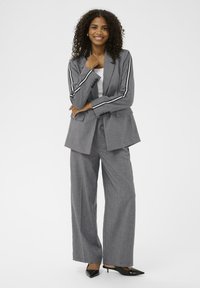 Sivi odijelo sa širokim nogavicama i kontrastnim bijelim prugama duž rukava. Materijal izgleda glatko, u kombinaciji s crnim cipelama s šiljastim vrhom.