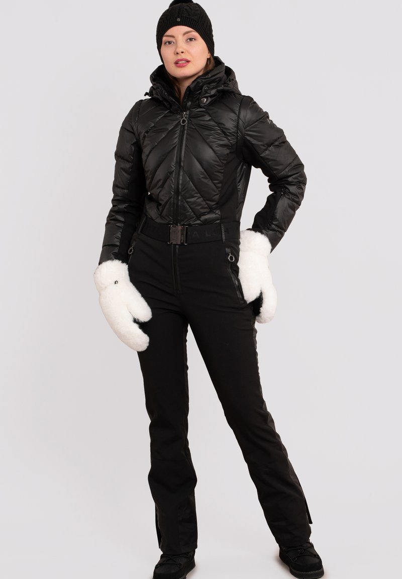 Vrouw in een zwarte pufferjack, zwarte broek, zwarte gebreide muts en grote witte wanten, staand met één hand op de heup tegen een effen achtergrond.