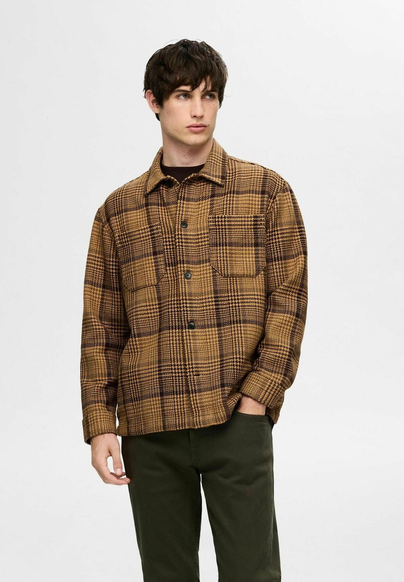 Brązowa koszula w kratę z kołnierzykiem, zapinana na guziki z przodu, z dwoma kieszeniami na piersiach. Wykonana z teksturowanej tkaniny. Połączona z ciemnozielonymi spodniami.