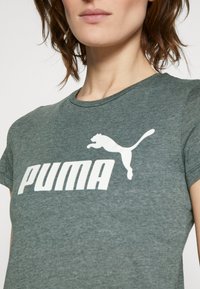 Grönmelerad T-shirt i mjukt tyg med en stor vit "PUMA"-logotyp och en hoppande puma-grafik. Klassisk rundhals och korta ärmar.