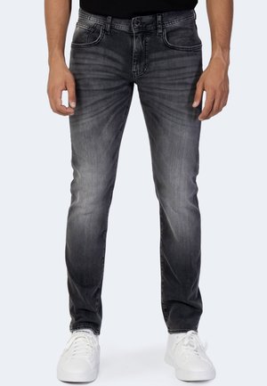 Hombre con jeans negros descoloridos de corte ajustado y zapatillas blancas, de pie con las manos relajadas a los lados frente a un fondo sencillo.