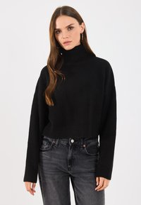 Schwarzer, kurzer Rollkragenpullover mit gerippter Textur und langen Ärmeln, kombiniert mit dunkelgrauen Jeans. Einfaches, minimalistisches Design.