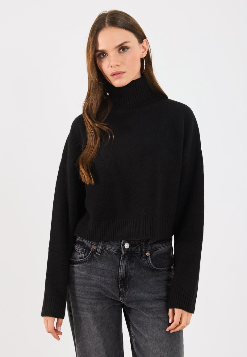 Schwarzer, kurzer Rollkragenpullover mit gerippter Textur und langen Ärmeln, kombiniert mit dunkelgrauen Jeans. Einfaches, minimalistisches Design.
