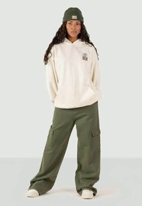 Giovane donna con felpa crema con toppano a tigre, pantaloni cargo oliva, sneakers bianche e berretto di maglia verde oliva, in piedi con le mani in tasca.