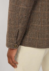 Bruine geruite blazer met een gestructureerde weving, voorzien van een knoopdetail op de manchetten, gecombineerd met lichtgekleurde ribfluwelen broek.