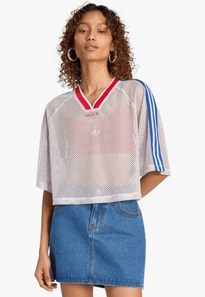 Donna che indossa un crop top in rete bianco Adidas con strisce blu e scollo rosso abbinato a una minigonna di jeans blu, in piedi su sfondo bianco.