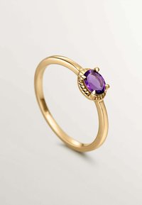 Anillo de oro con una gema ovalada de color púrpura, montada en un engaste con garras y con un detalle texturizado alrededor de la piedra, diseño de banda suave.