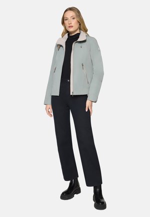 Woman standing with hands relaxed, wearing light gray zip-up jacket, black turtleneck, black wide-leg pants, and black ankle boots.