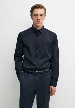 Ung mand iført en tætsiddende sort skjorte med knapper og matchende sorte bukser med sort bælte mod en ensfarvet lys baggrund.