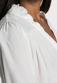 Blusa bianca con scollo arricciato, tessuto morbido e maniche a palloncino, che presenta sottili dettagli stropicciati e una vestibilità rilassata.